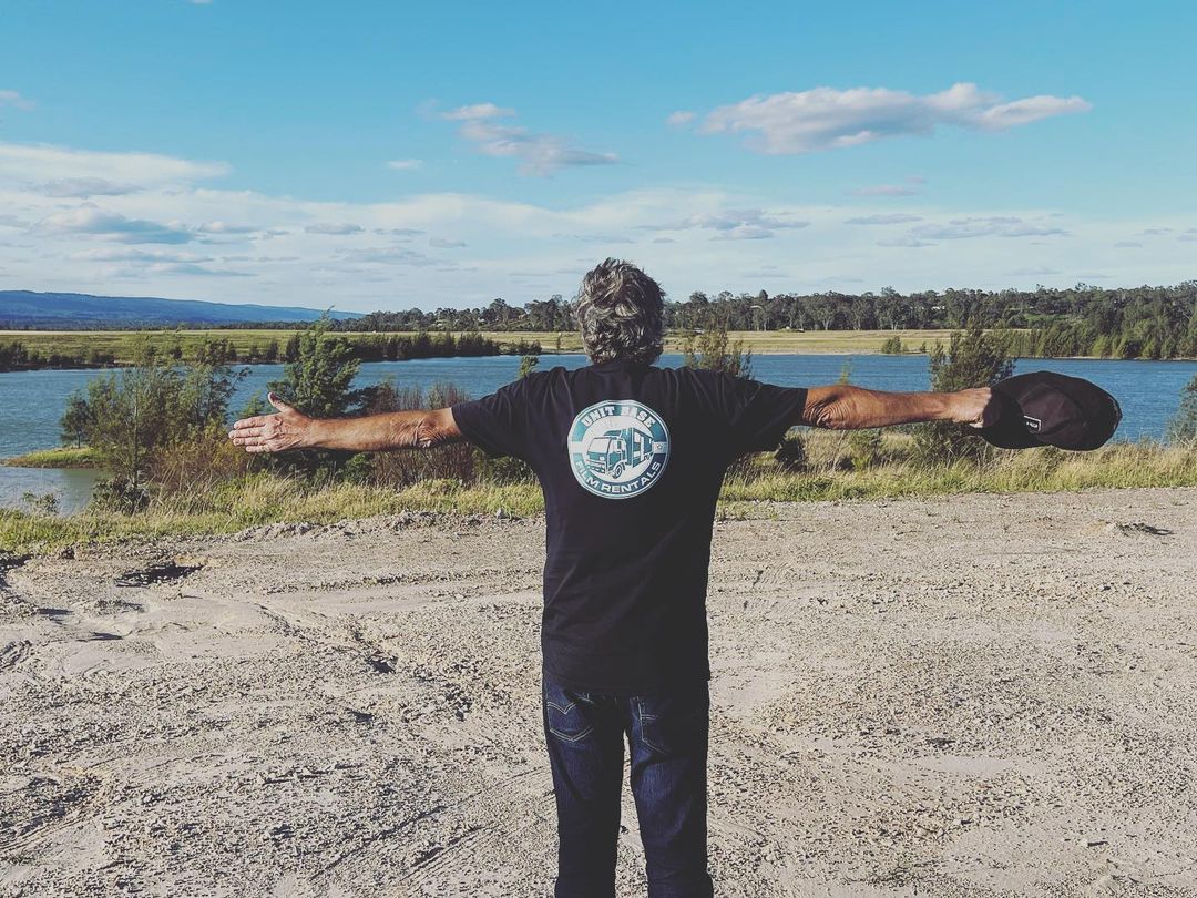 Person stands with arms outstretched near a body of water under a bright sky, enjoying the natural scenery, wearing a dark t-shirt.