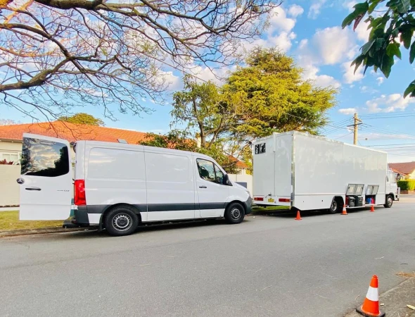 Two white service vans parked on a street next to a suburban home, tree branches overhead, under a blue and partly cloudy sky.