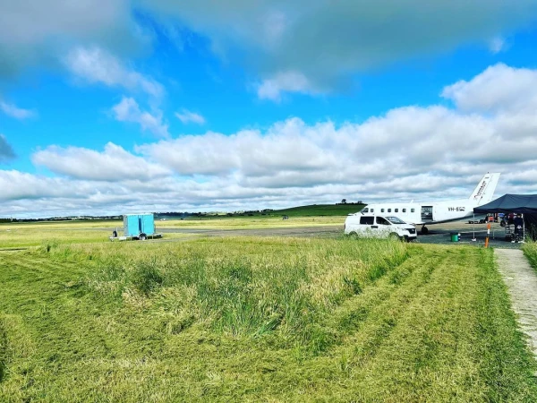 White single-engine airplane parked on a rural airstrip with a white van, blue trailer, and grassy field under a cloudy blue sky.