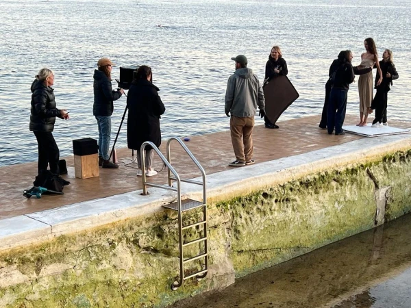 Outdoor fashion photoshoot on a pier featuring models in flowing dresses with a photography crew and tranquil ocean backdrop.