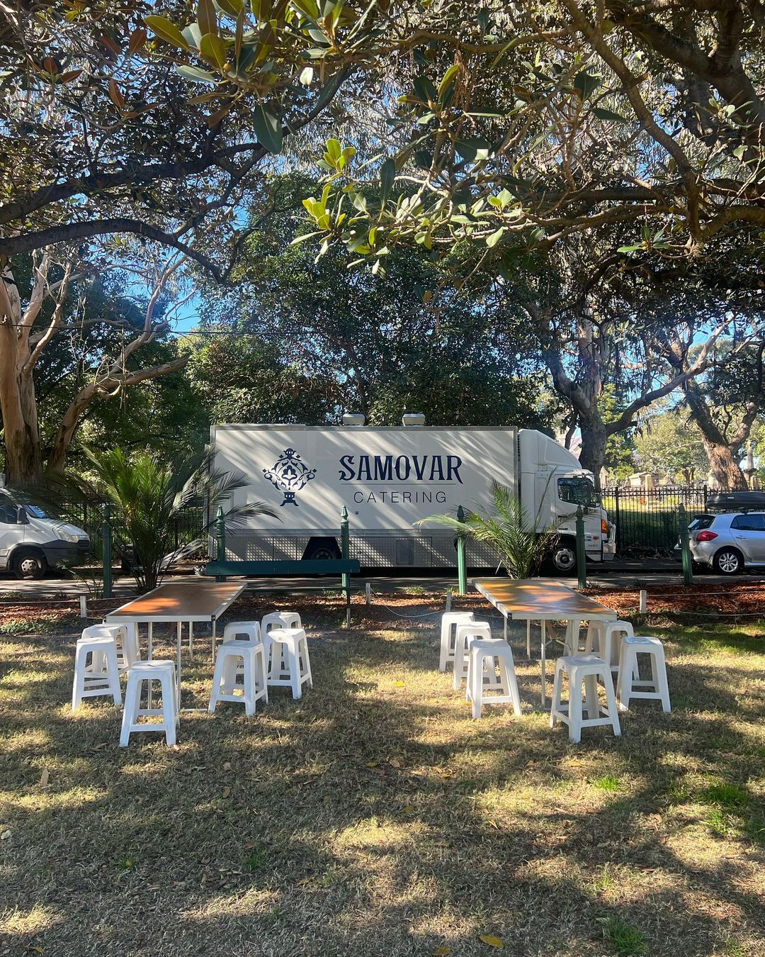 Outdoor catering setup with a Samovar Catering truck, tables, and white chairs arranged on a lawn under a tree.