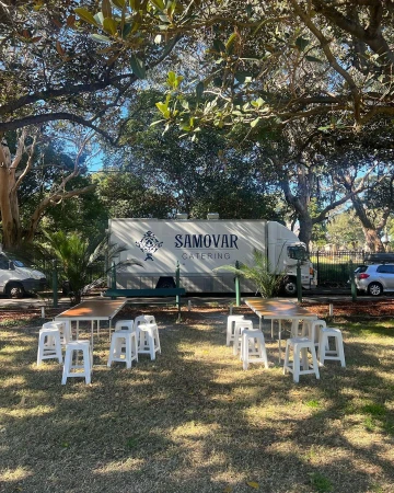 Outdoor catering setup with a Samovar Catering truck, tables, and white chairs arranged on a lawn under a tree.
