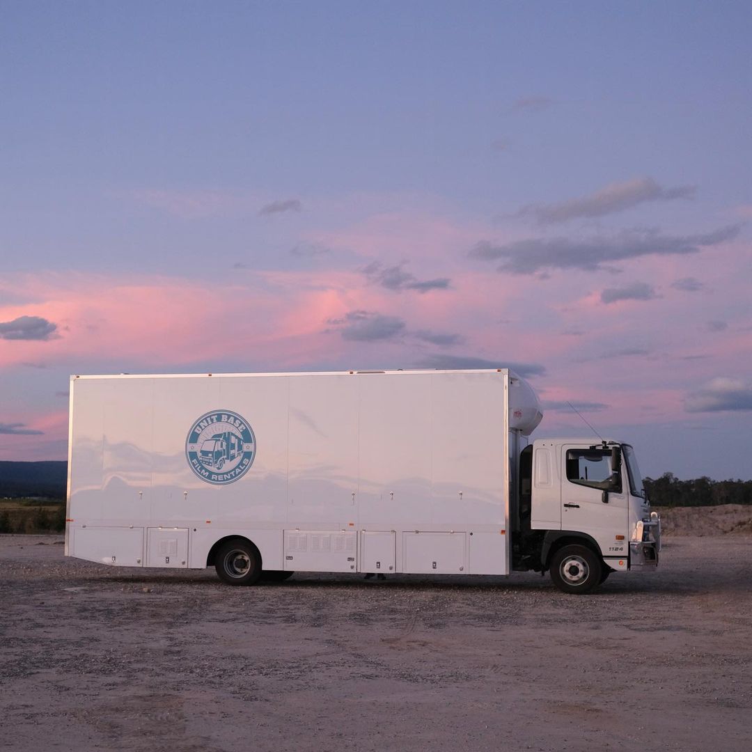 A white box truck is parked in a gravel lot, under a pastel sky with soft clouds, near shrubbery and small trees in the background.