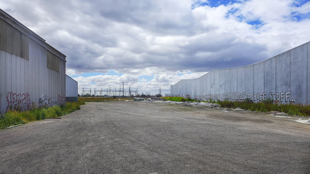 Industrial landscape featuring large concrete walls covered with graffiti, set against a cloudy sky with distant power lines and gravel ground.