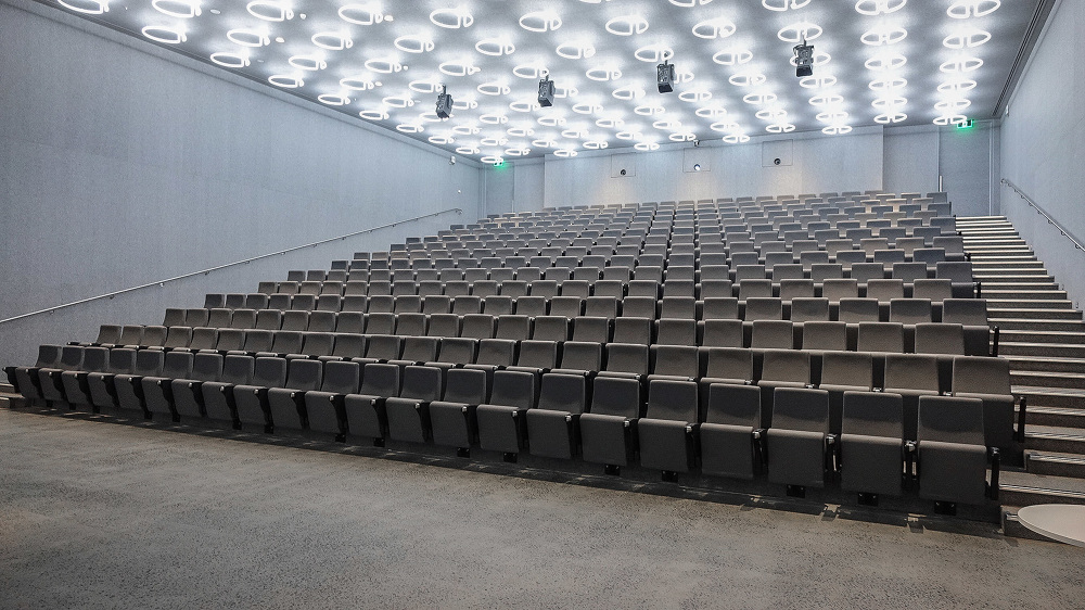 Interior view of an auditorium with rows of gray upholstered seating, a concrete floor and walls, and overhead lighting rings.