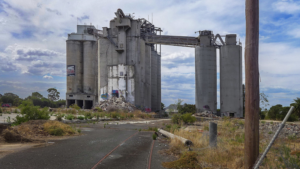 Wide shot of an abandoned concrete industrial complex with multiple silos and connecting structures under a cloudy sky in an overgrown area.