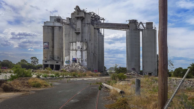 Wide shot of an abandoned concrete industrial complex with multiple silos and connecting structures under a cloudy sky in an overgrown area.