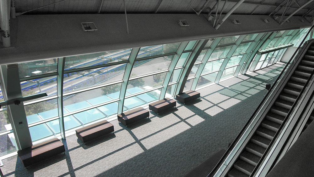 Interior view from above shows airport terminal area with windows, benches, escalator, and carpeted floor.