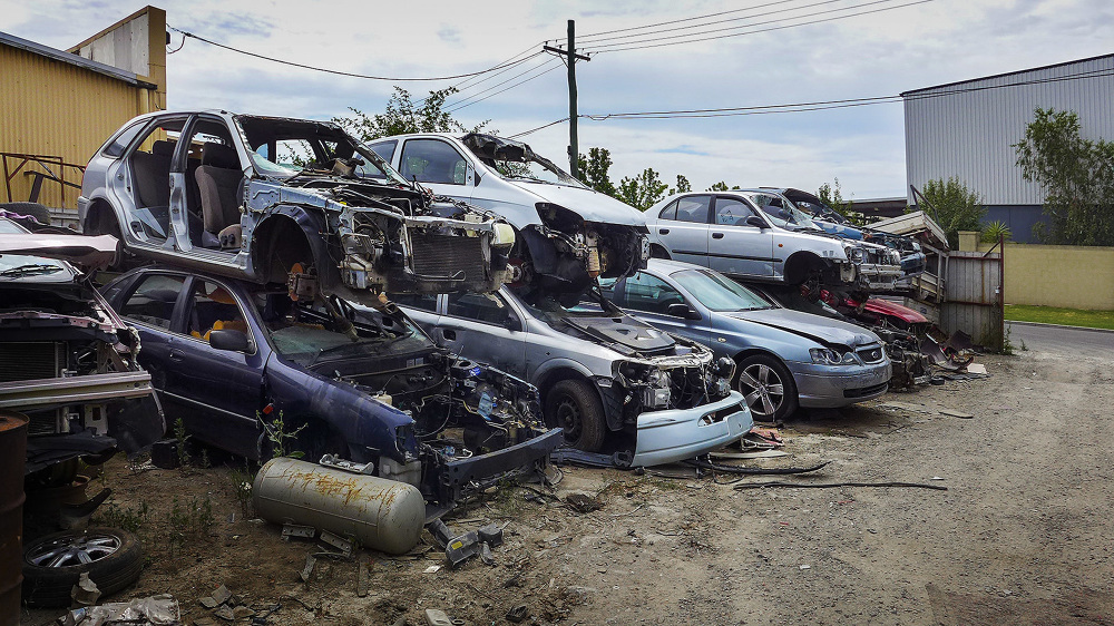 Outdoor image of a junkyard with a collection of wrecked cars and auto parts, under an overcast sky, and various industrial buildings.