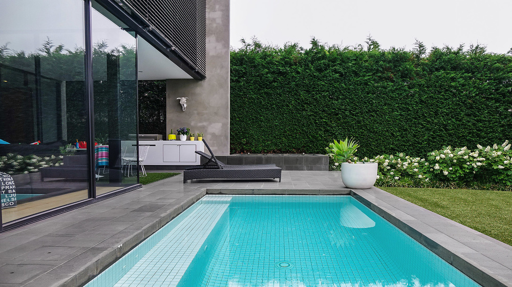 Modern outdoor living space featuring a turquoise swimming pool, lounge chair, manicured greenery, and a contemporary building design.