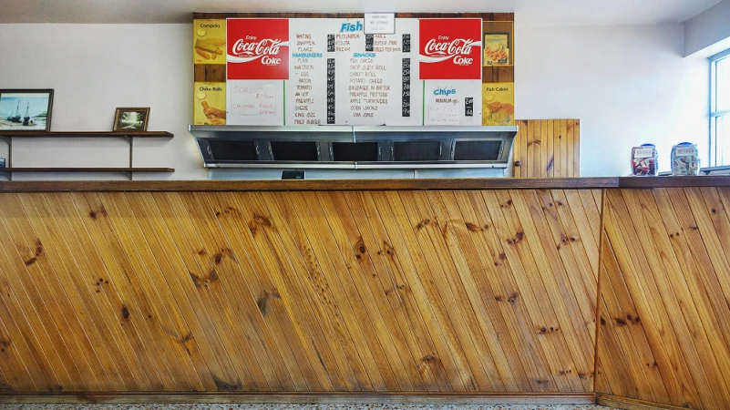 Interior view of a vintage fish and chip shop with a wooden counter, a menu board featuring Coca-Cola ads, and handwritten food pricing.