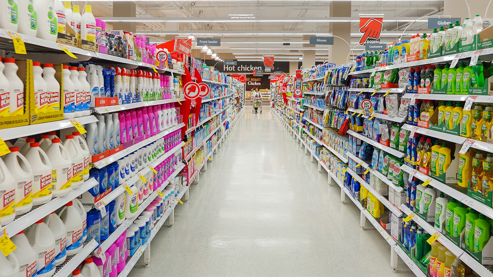 Brightly lit supermarket aisle with shelves stocked with cleaning supplies and household goods, offering a variety of product choices.