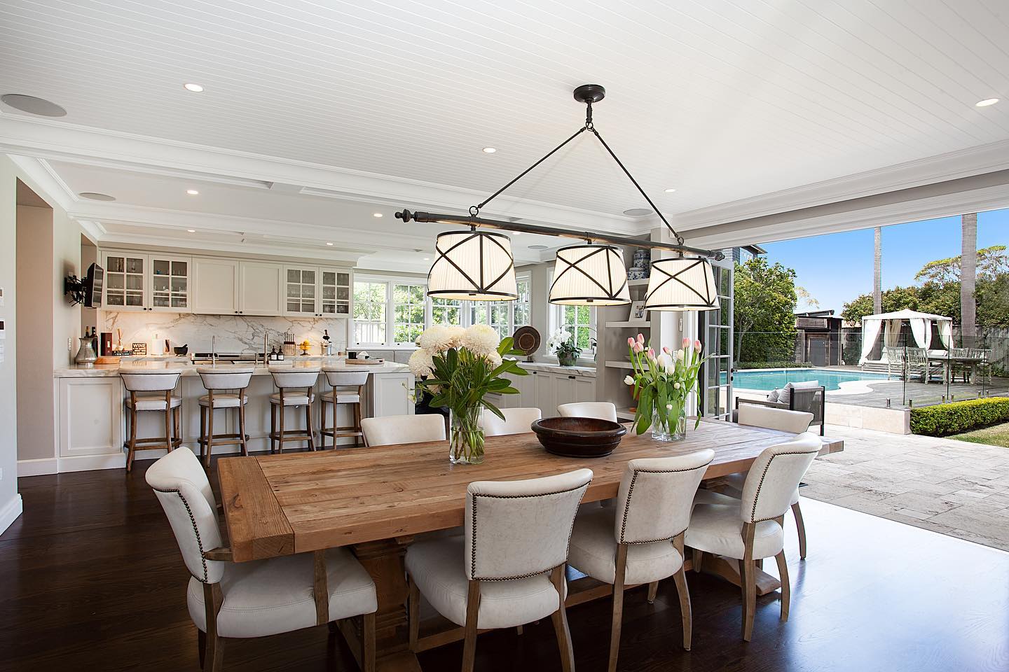 Bright, spacious kitchen and dining room with a wooden table, white chairs, and a view of the pool area in the background.