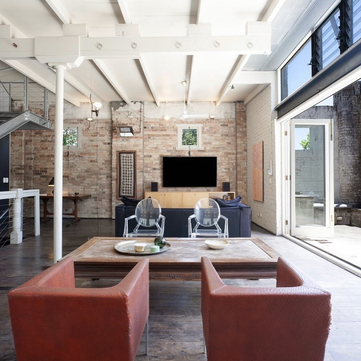 Living room interior featuring exposed brick wall, modern furniture including transparent chairs, and large glass sliding doors to exterior.