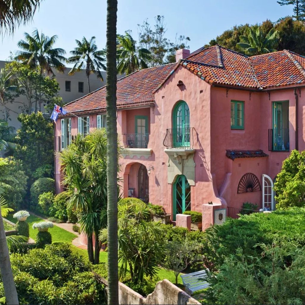 Exterior shot of a two-story pink house with green trim, featuring a red tile roof, arched windows, and a well-maintained garden.