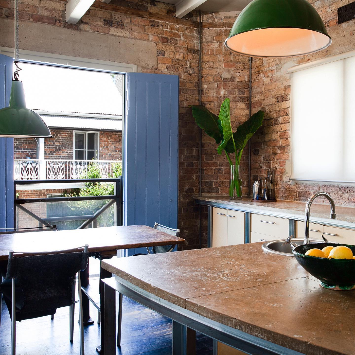 Modern kitchen interior with brick walls, featuring blue doors to the outside, stainless steel counters, lemons, and a hanging green light.
