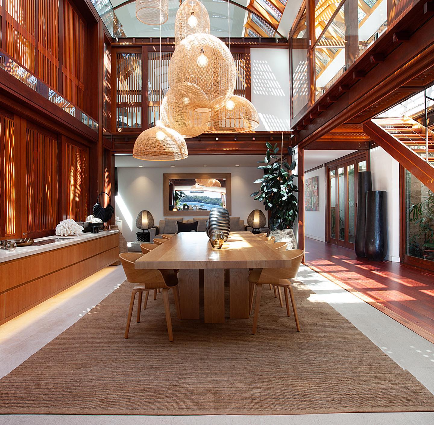 View of a modern dining room with wooden table and chairs under woven pendant lights in a high-ceiling room with wood accents and a large rug.