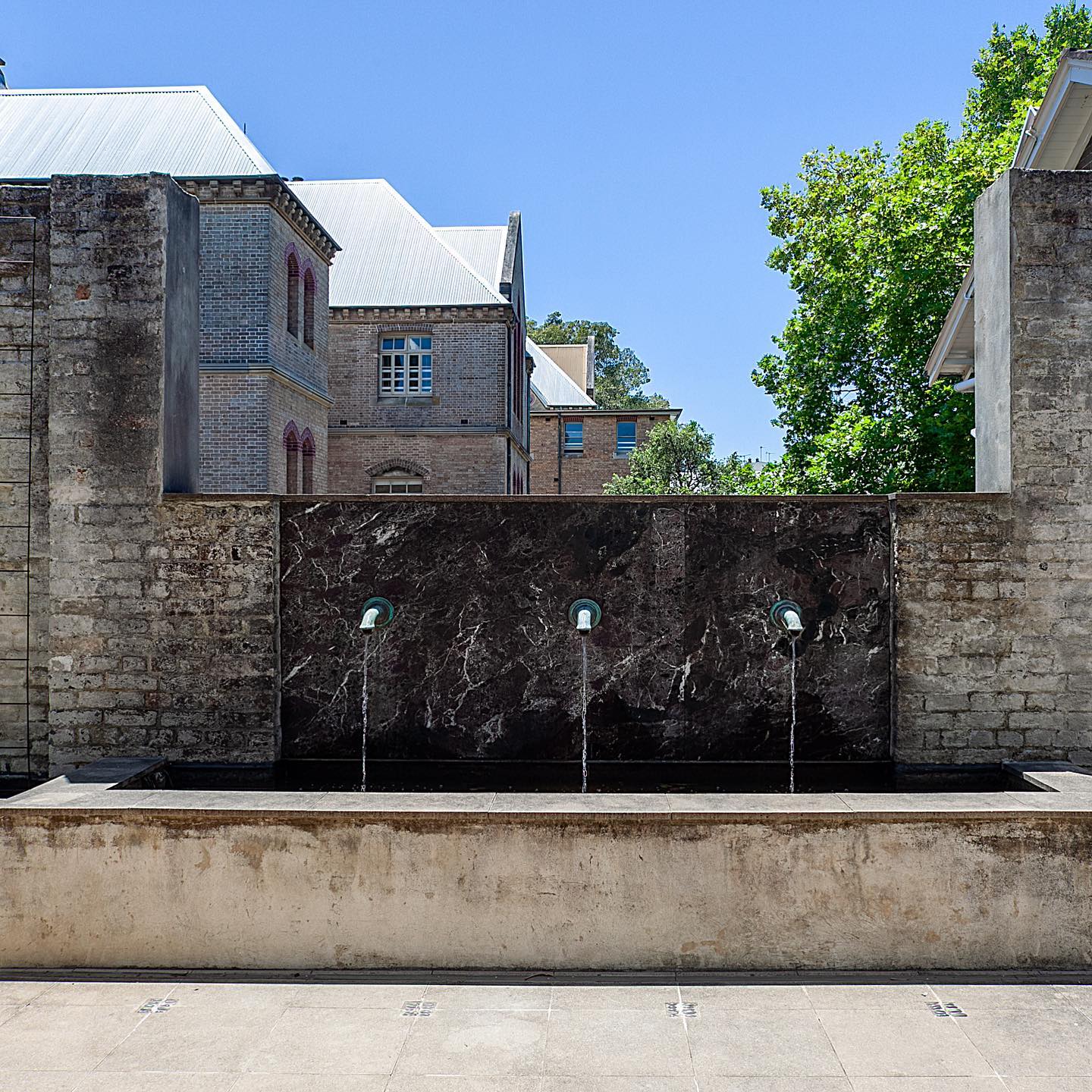 Outdoor water fountain with three spouts set against a dark marble background, flanked by aged brick walls, and building in the distance.