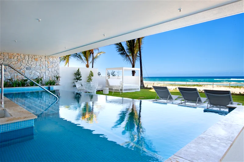 View of pool merging with ocean at a resort, showcasing crystal-clear water, lounge chairs, and tropical palm trees under a blue sky.