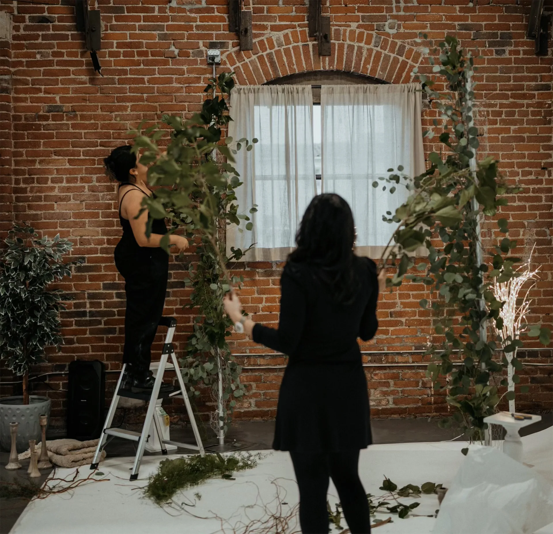 Two women arranging leafy green branches on an archway against a brick wall backdrop, creating a natural and elegant floral display.
