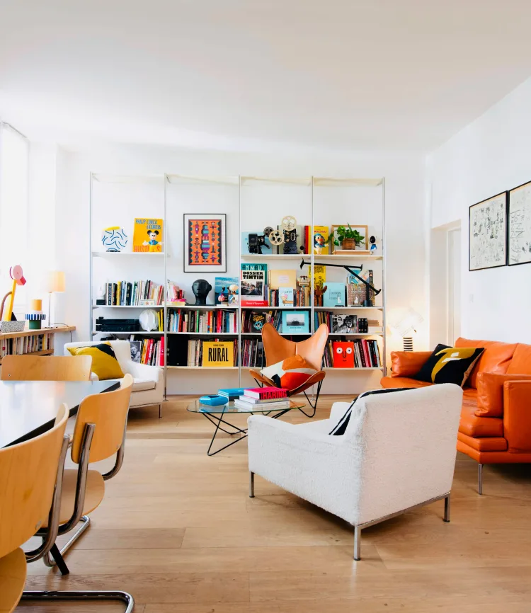 Bright living room with a bookshelf, orange sofa, white armchairs, a tan butterfly chair, and light wood floors in natural light.