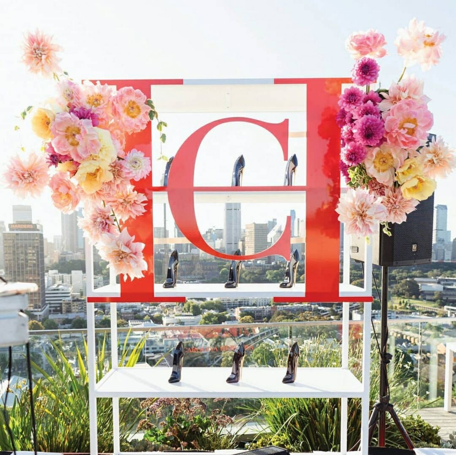 Fashion display with black high-heeled shoes arranged on a tiered red and white shelf, accented by a floral arrangement and city view.