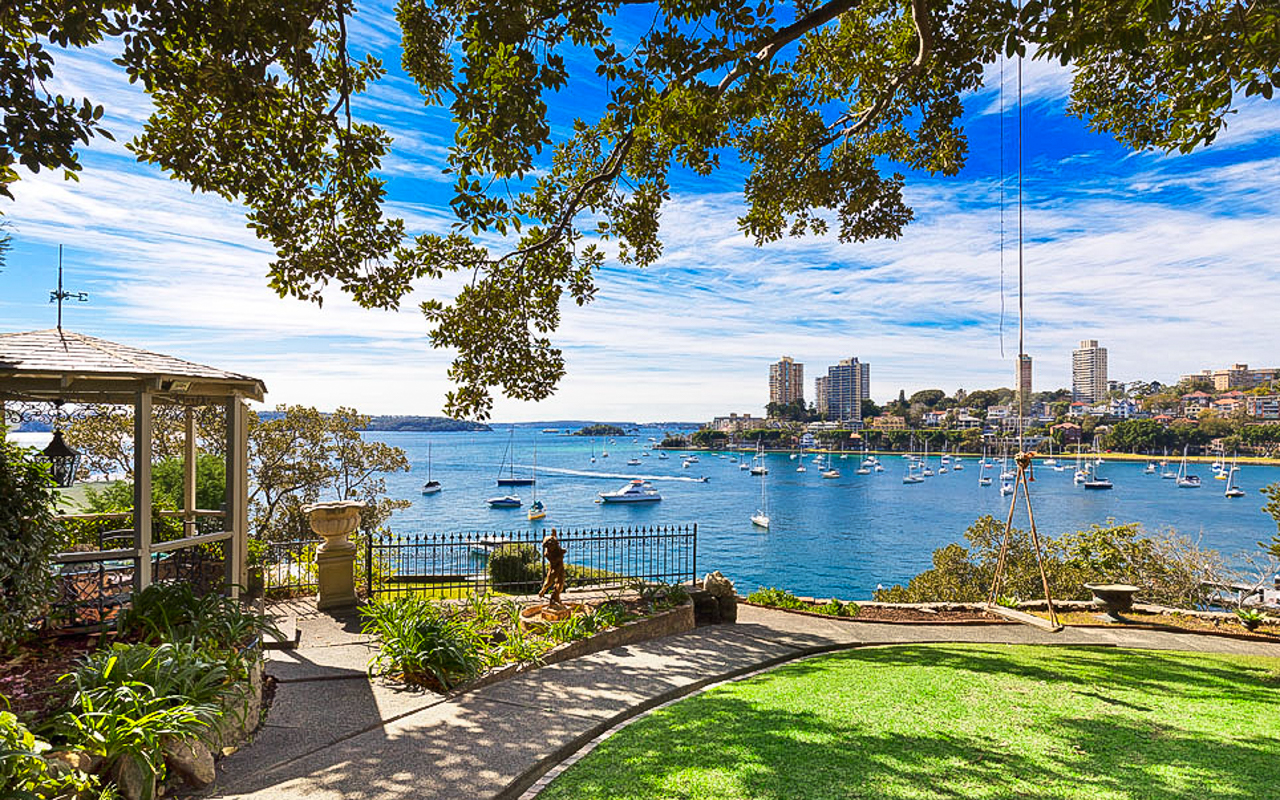 Scenic view of a harbor with sail boats, a gazebo, and buildings in the background, captured from a grassy lawn on a sunny day.