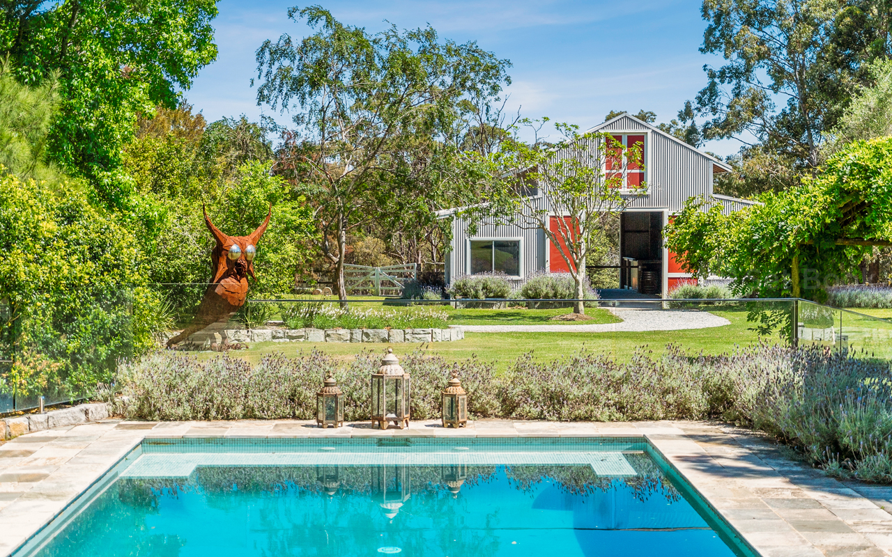 Outdoor swimming pool with clear blue water, stone tile surround, a metal sculpture, and a large barn-style house in the background.