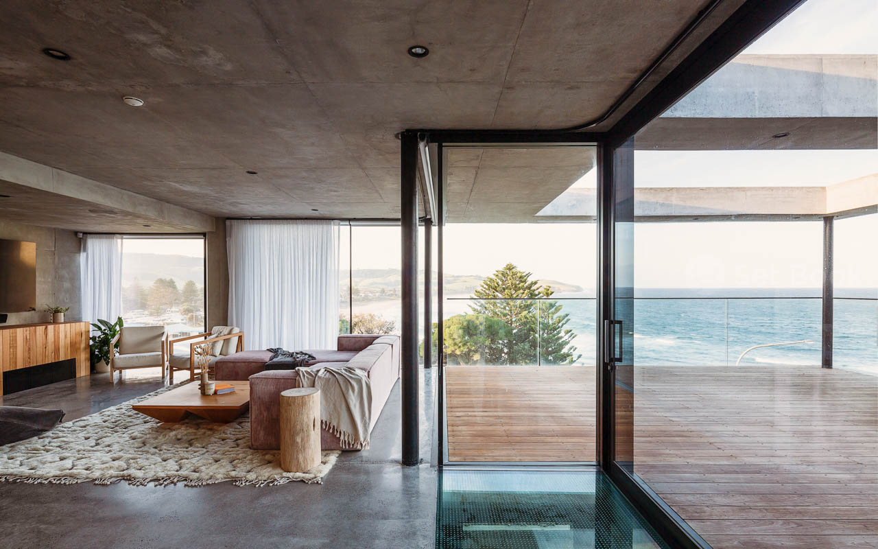 Modern home interior features a concrete ceiling and floor, large windows overlooking the ocean, and stylish furniture arrangement.