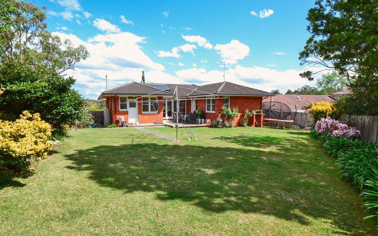 Exterior view of a single-story red brick house with a dark roof and a large green lawn, taken on a sunny day with blue skies.