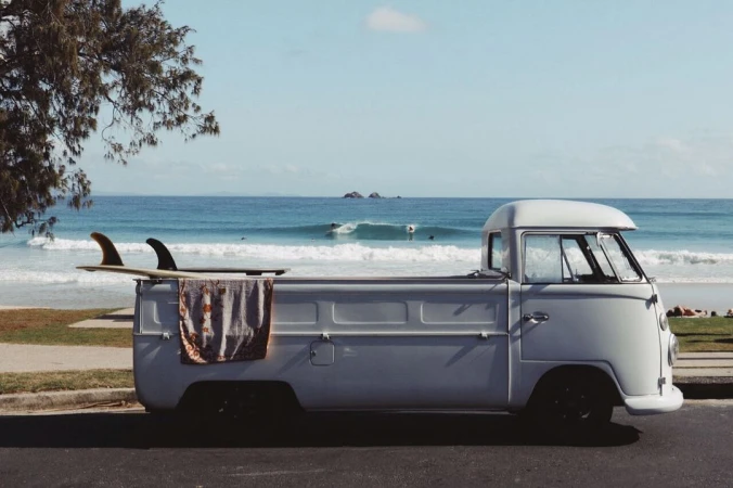 Vintage camper van parked near a beach with surfboards, blue ocean waves, clear sky, and surfers riding waves in the background.