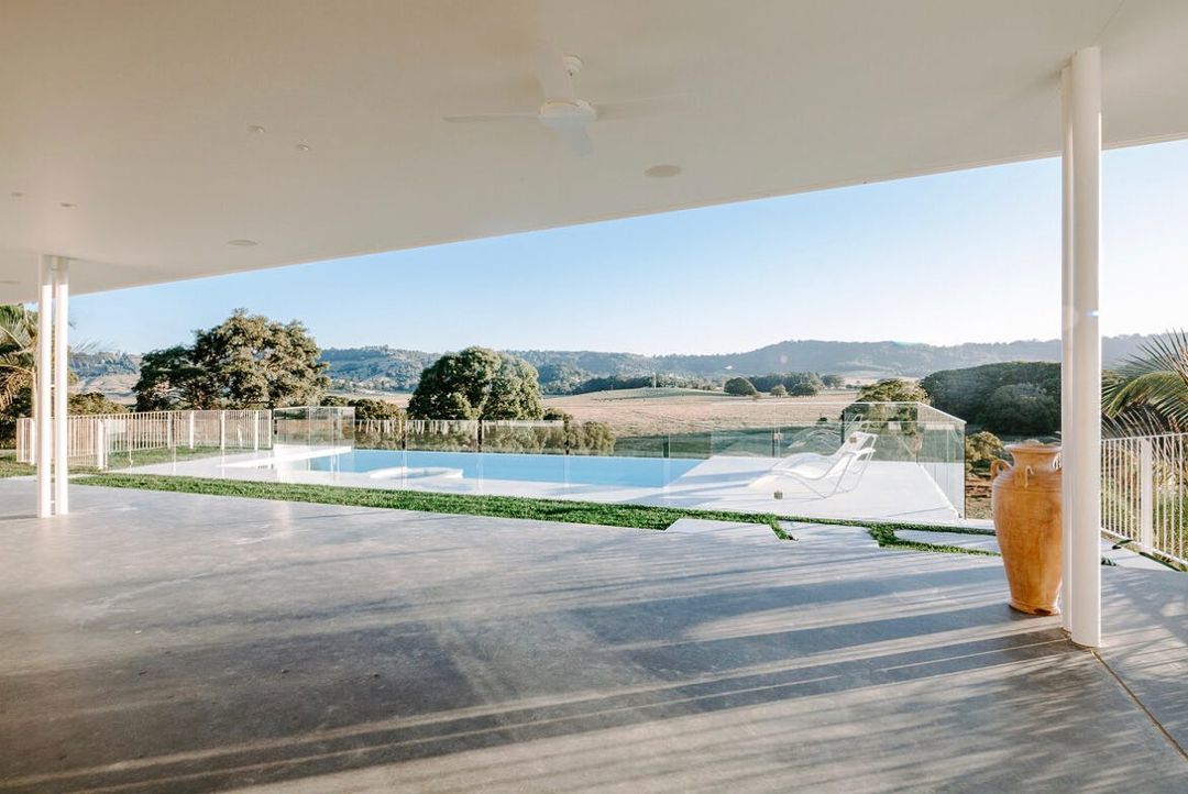 Outdoor view of a modern home with a swimming pool, deck chairs, and a vase overlooking a lush green landscape under a clear blue sky.