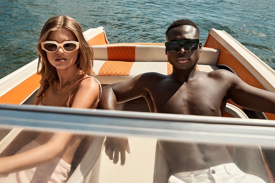 Two attractive people, an adult female and an adult male, wear sunglasses on a boat with orange trim on the water on a sunny day.