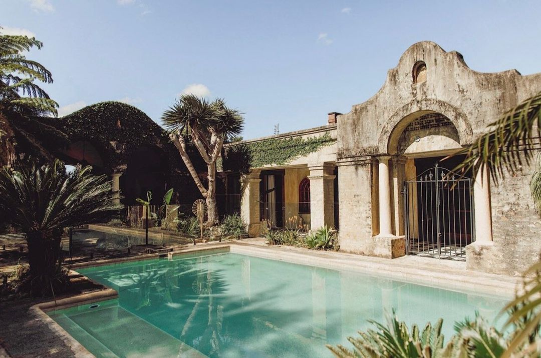 Exterior view of a Spanish Colonial Revival house with a swimming pool, stone walls, and lush greenery on a sunny day, creating a serene scene.
