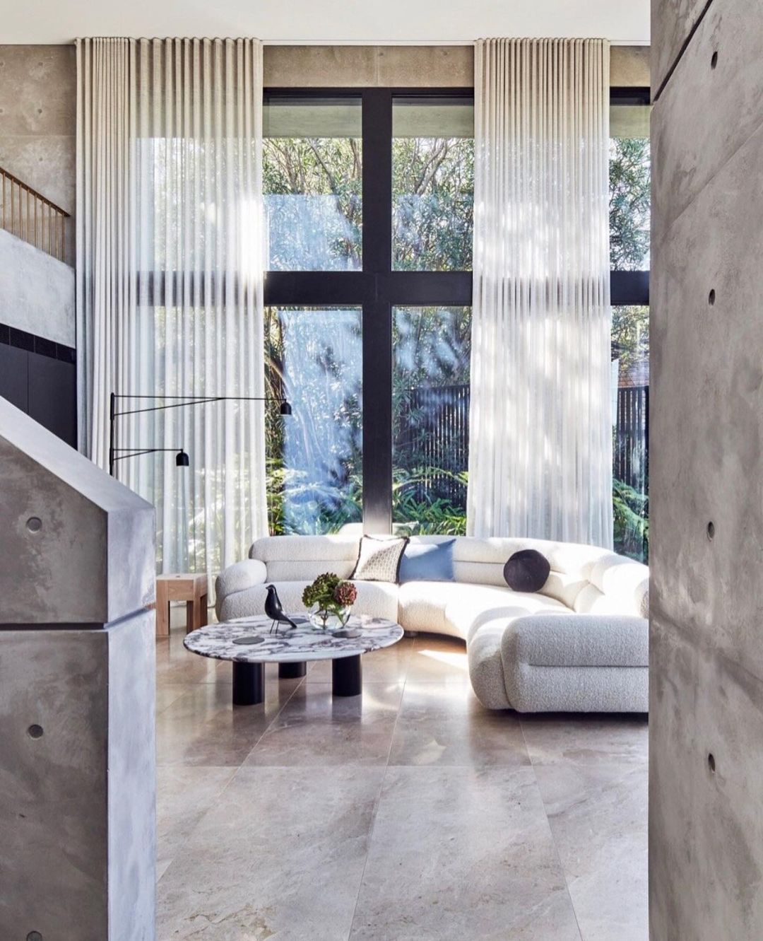 Interior view of a modern living room featuring a white rounded sectional sofa, floor-to-ceiling windows, and a marble coffee table.