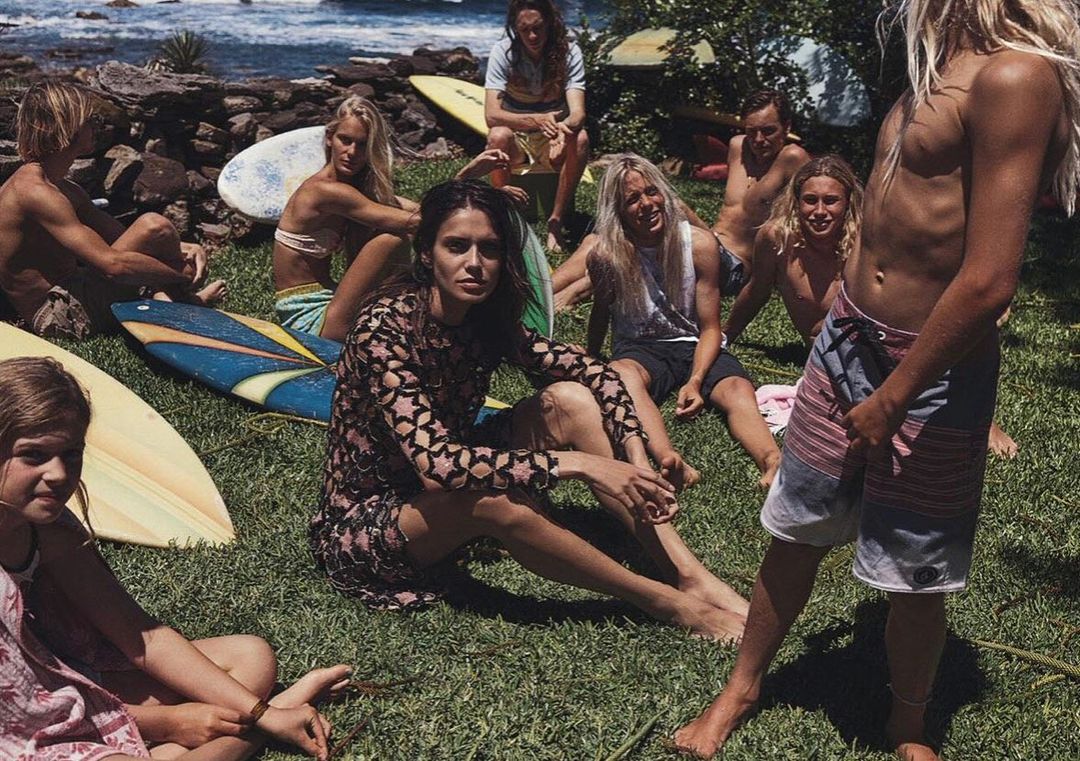 Group of young adults gather on a grassy lawn near the ocean with surfboards, enjoying a casual, sunny day together in a relaxed setting.
