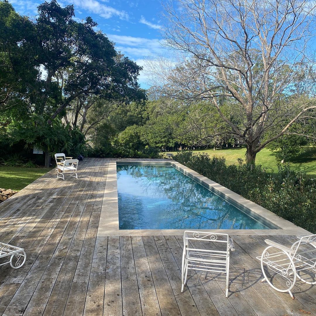 Outdoor swimming pool surrounded by a wooden deck and lush greenery under a bright blue sky with trees. Summer setting with calming water.