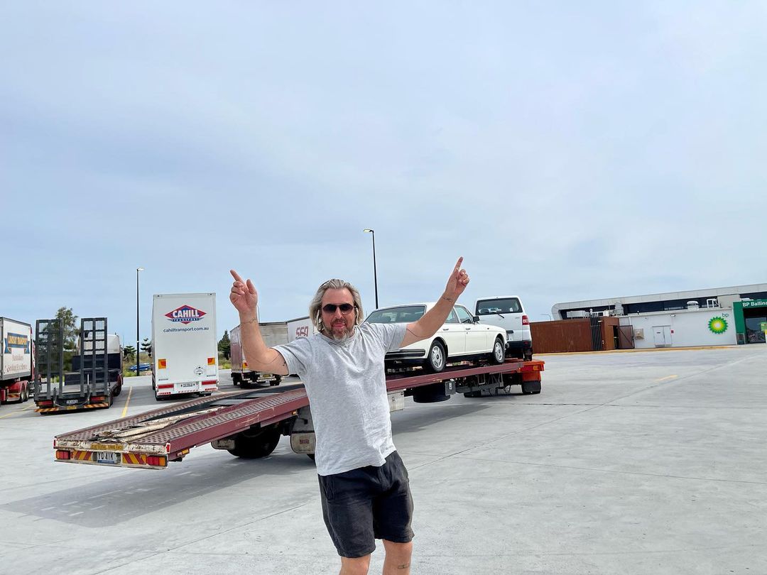 Adult male with arms raised stands in front of a car on a flatbed truck on a paved lot, with other trucks and buildings nearby.