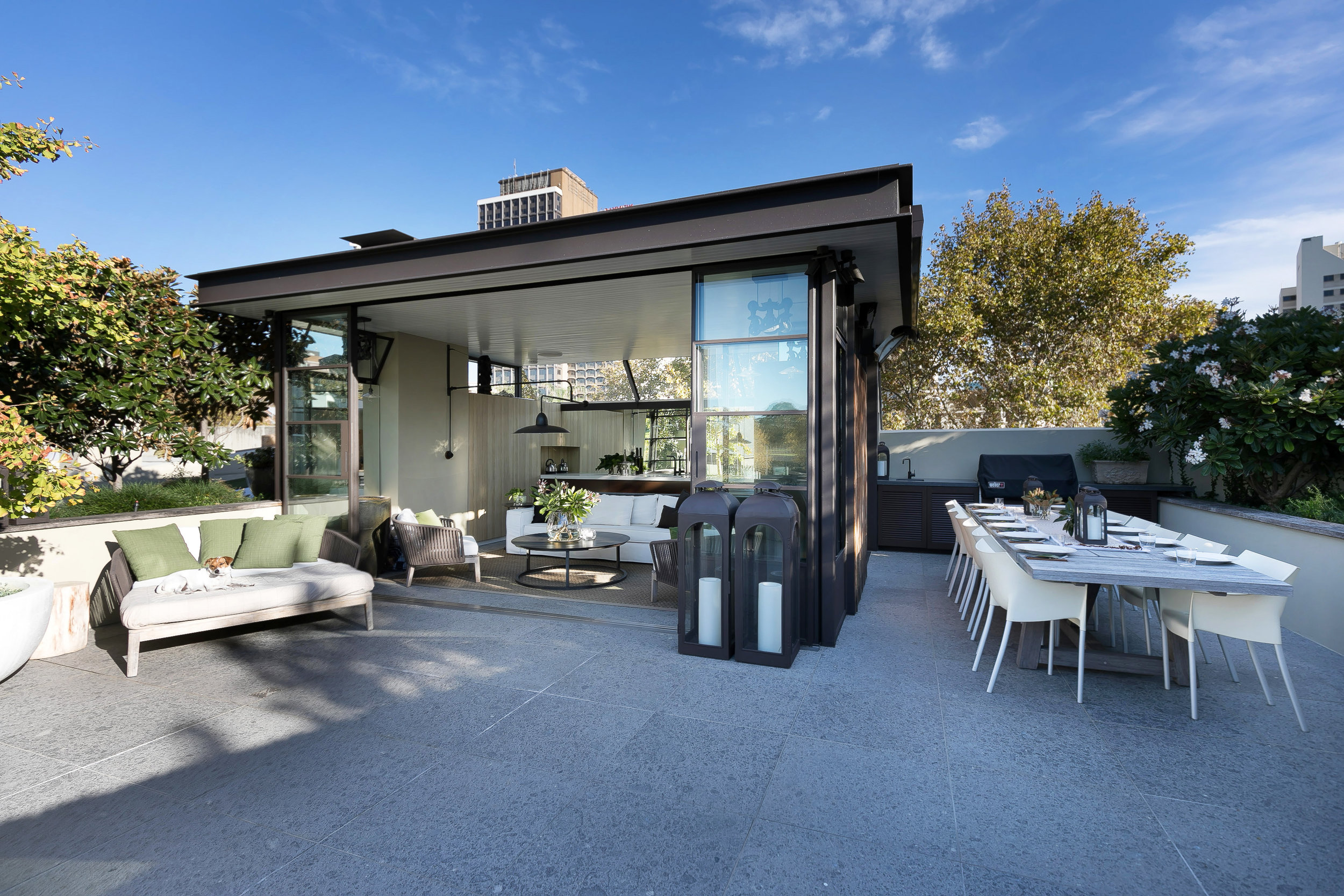 Outdoor patio with dining table set for 10, modern lounge set with green pillows, and exterior lighting. Sunny blue sky overhead.