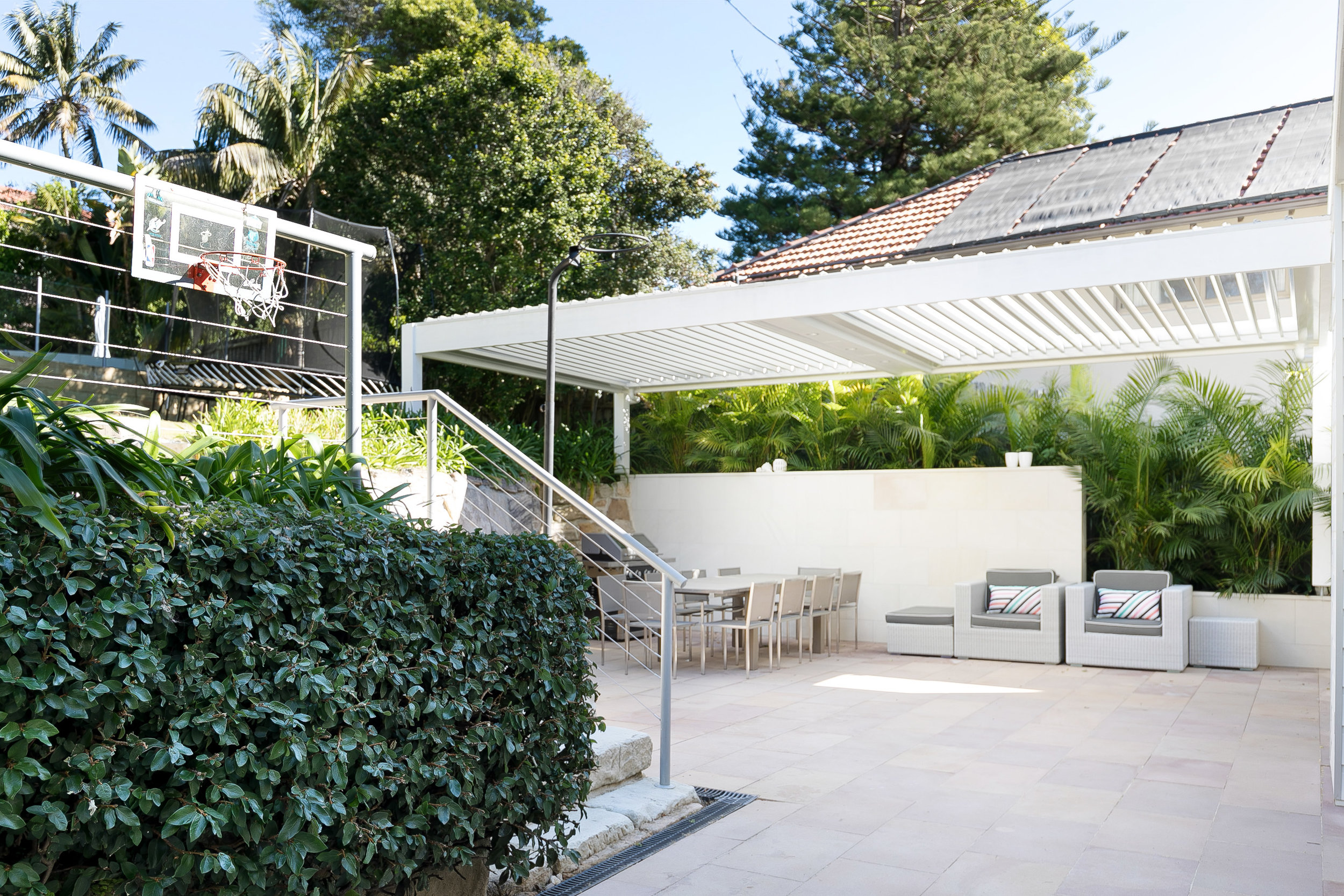 Outdoor patio with modern furniture, a dining table set, and lush greenery under a white pergola, offering a luxurious backyard space.
