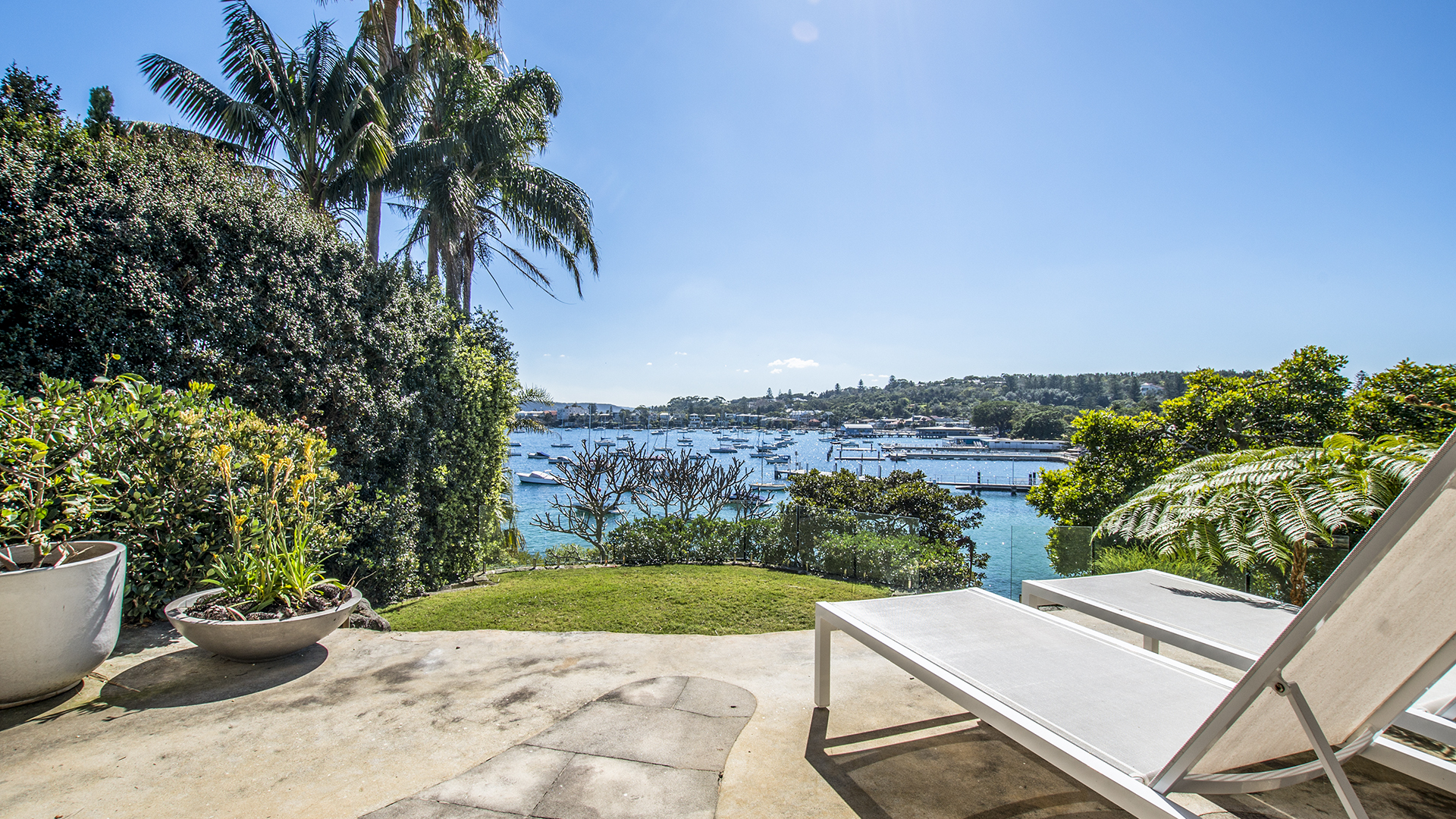 Outdoor patio with lounge chairs overlooking a serene harbour with boats and lush greenery, showcasing a tranquil waterside setting.