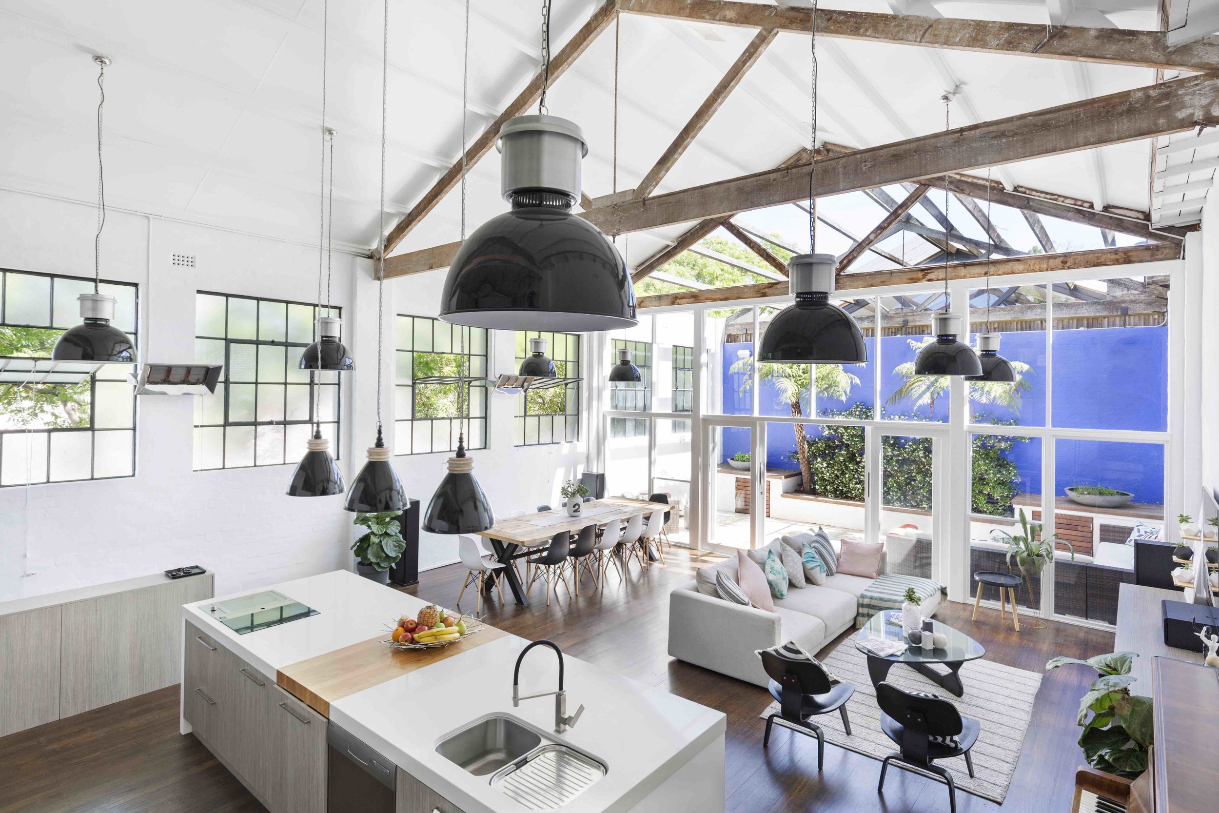 Brightly lit, high-ceilinged open-plan modern apartment interior with a kitchen island, dining table, sitting area and black pendant lights.