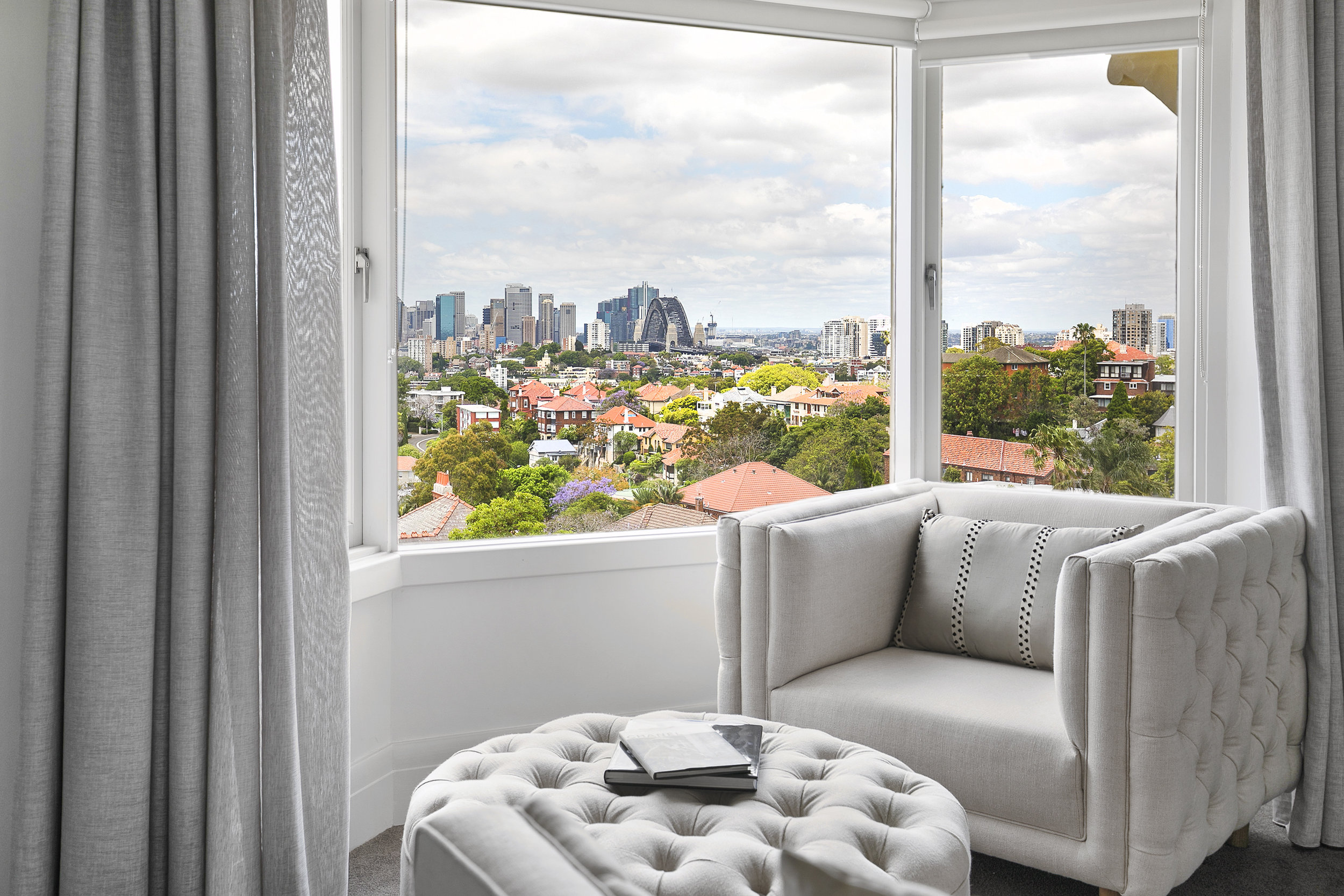 Living room interior featuring a cream-colored armchair and tufted ottoman, with a city skyline view visible through the large window.