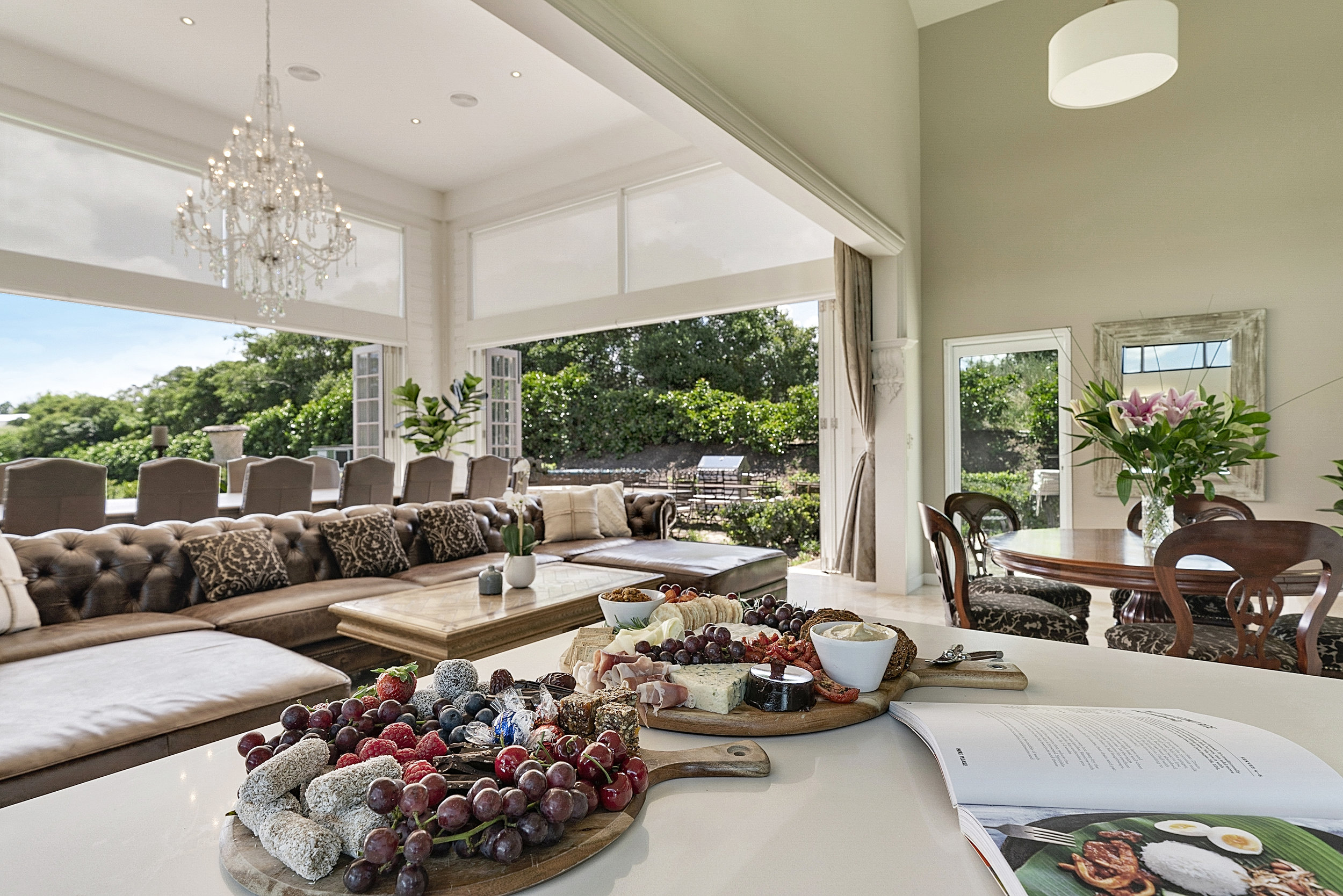 Interior of a well-lit home with charcuterie boards on a kitchen counter, a chandelier, and large windows with greenery outside.