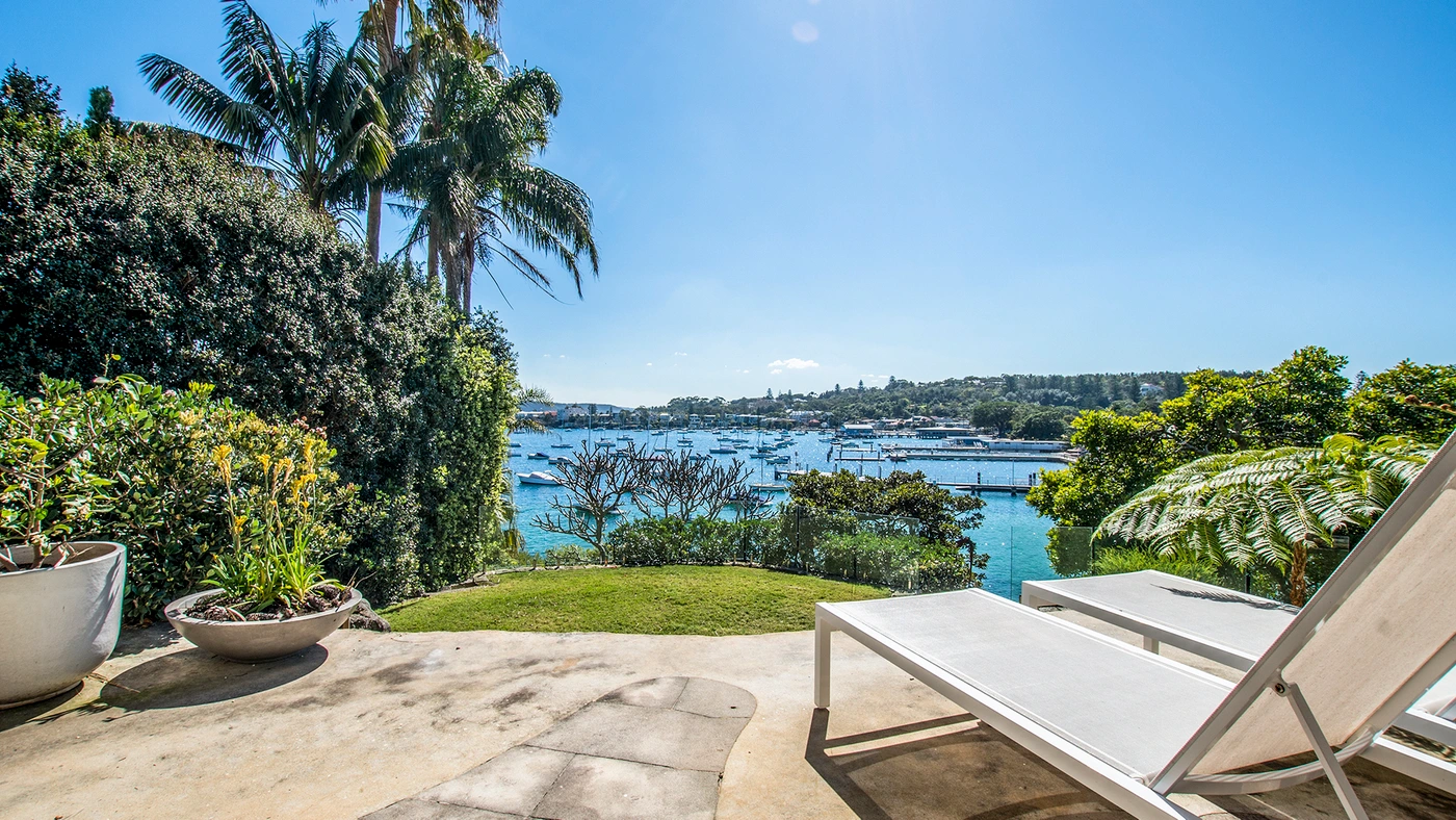 Sunny waterfront terrace in Sydney featuring two white sun loungers, lush palm trees, and a view of docked boats on blue water.
