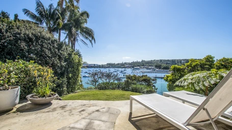 Sunny waterfront terrace in Sydney featuring two white sun loungers, lush palm trees, and a view of docked boats on blue water.