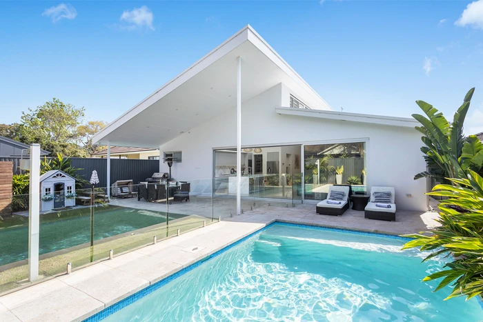 Modern white house exterior with a sloping roof, backyard swimming pool, and outdoor lounging area under a clear sky.