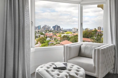 A beige armchair and tufted ottoman with books placed in front of a large bay window overlooking Sydney city skyline.