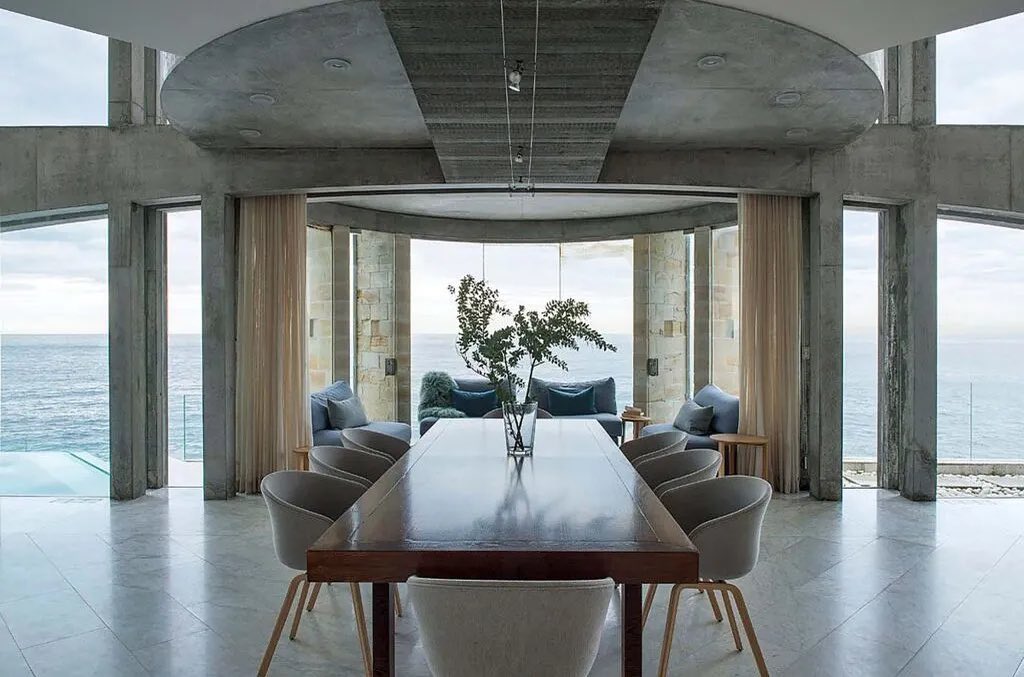 Modern dining room interior with a long wooden table and cushioned chairs, featuring ocean view through large windows in a concrete structure.