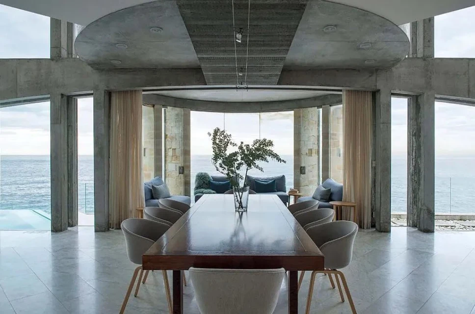 Modern dining room interior with a long wooden table and cushioned chairs, featuring ocean view through large windows in a concrete structure.